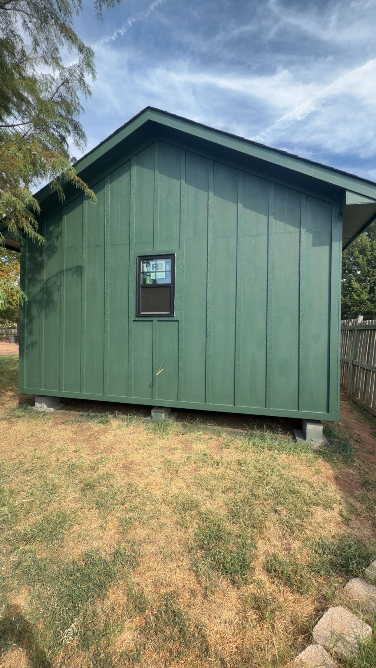 Dark green gabled shed with vertical siding and a single window elevated on concrete blocks.