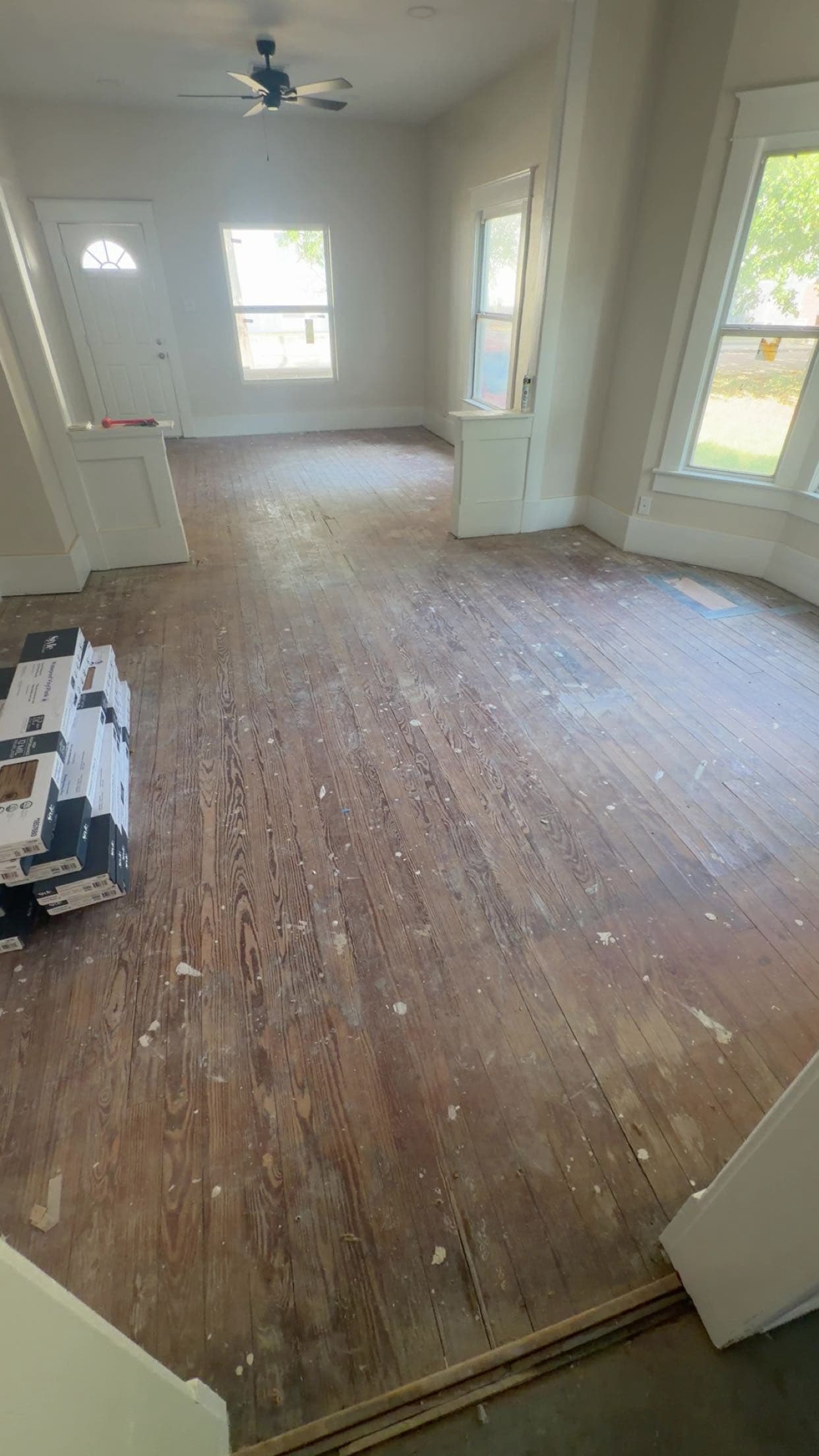 Empty room undergoing renovation with worn hardwood floors, paint splatters, and stacks of new flooring.