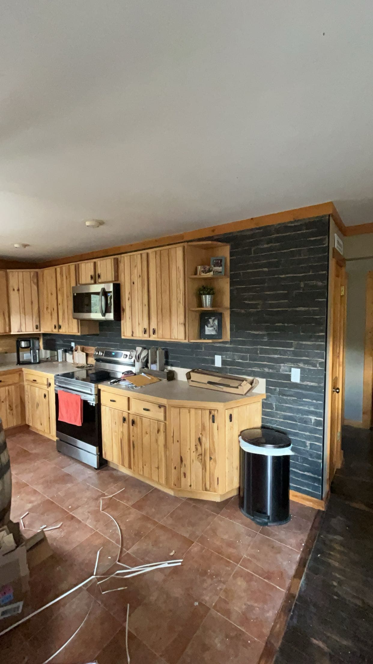 Light wood kitchen cabinets, dark brick accent wall, stainless steel appliances, and brown tile floor.